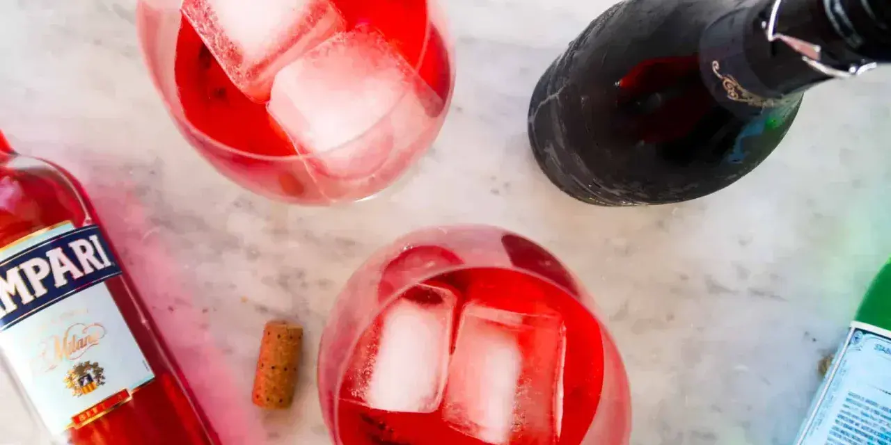 Dwa orzeźwiające Campari Spritz z lodem, butelka Campari i butelka prosecco na marmurowym blacie.