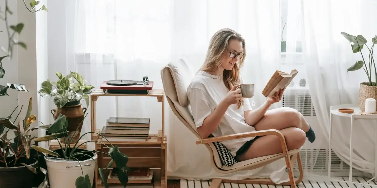 Mujer con gafas disfruta de un libro y una taza en su rincón de lectura, rodeada de plantas y luz natural.