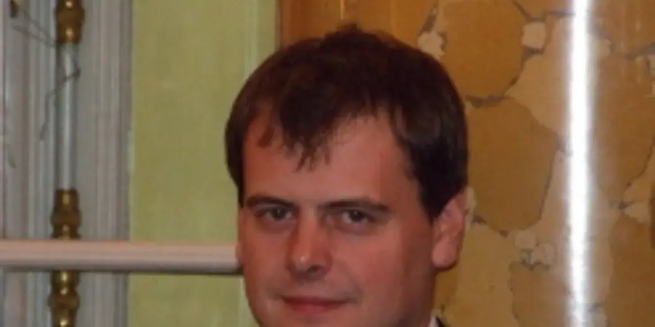 Tomasz Bartoś, zamyślony, w eleganckim stroju, na tle ozdobnej ściany.