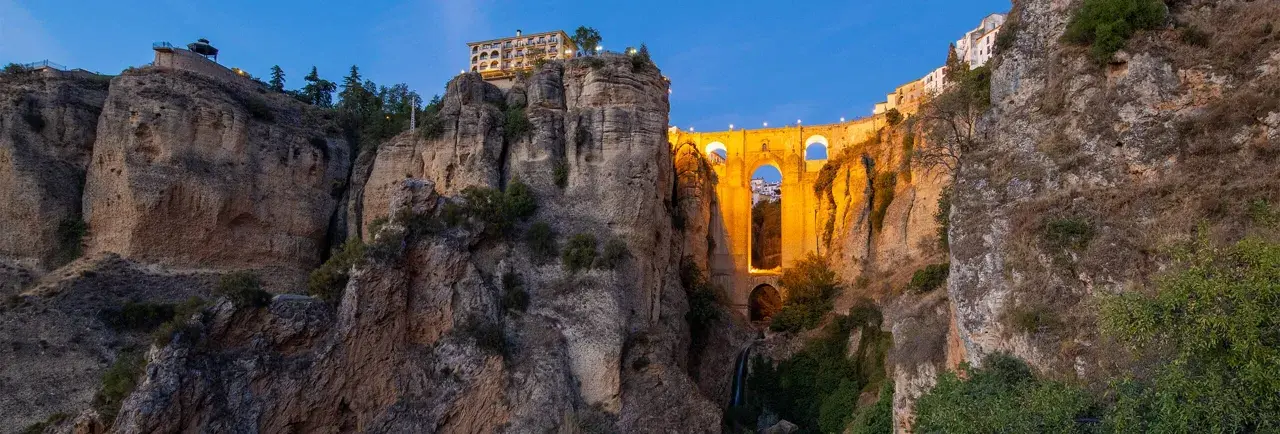A dramatic **Ronda bridge view point** at dusk, with the illuminated bridge spanning a deep gorge and buildings perched on the cliff edge.