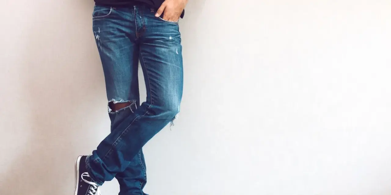 Mężczyzna w modnych jeansach z dziurami na kolanie, czarnych trampkach i czarnej koszulce, opiera się o ścianę.