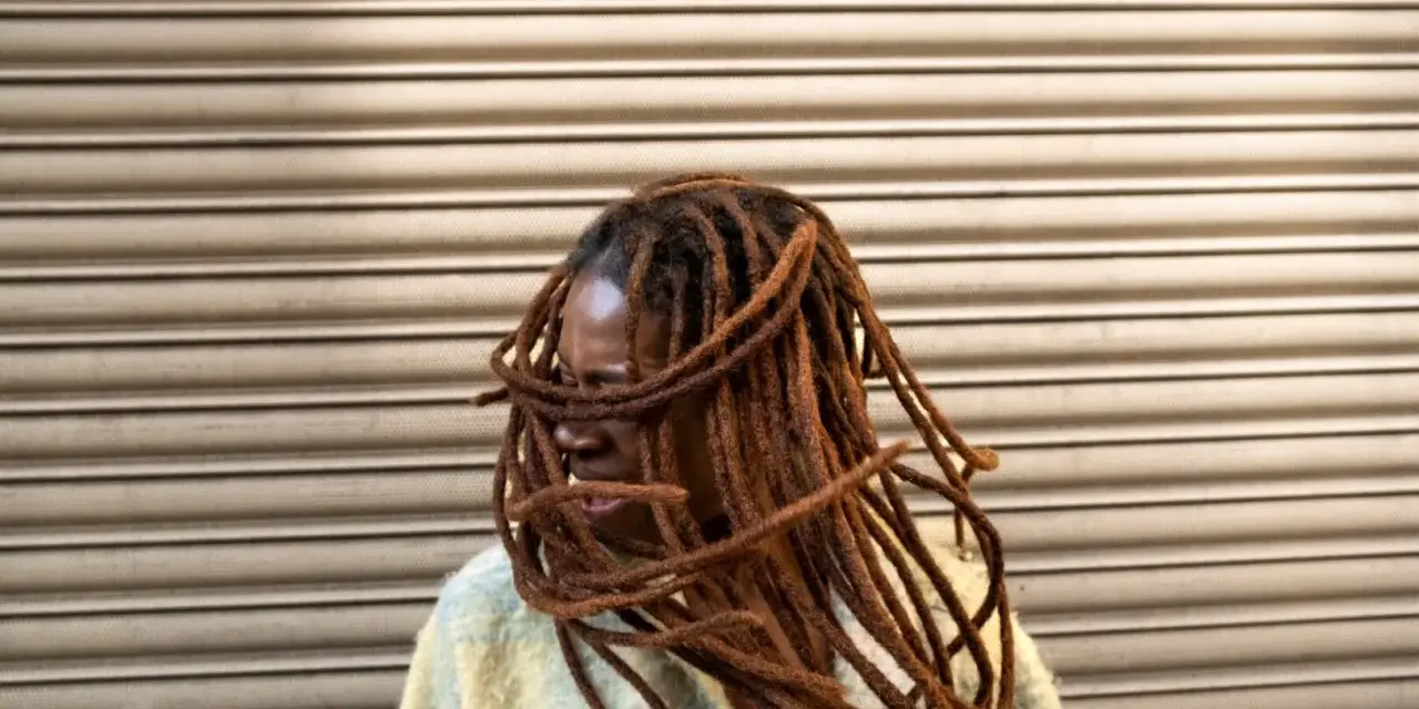 Donna con lunghe treccine africane che le incorniciano il viso, con uno sfondo a righe orizzontali.