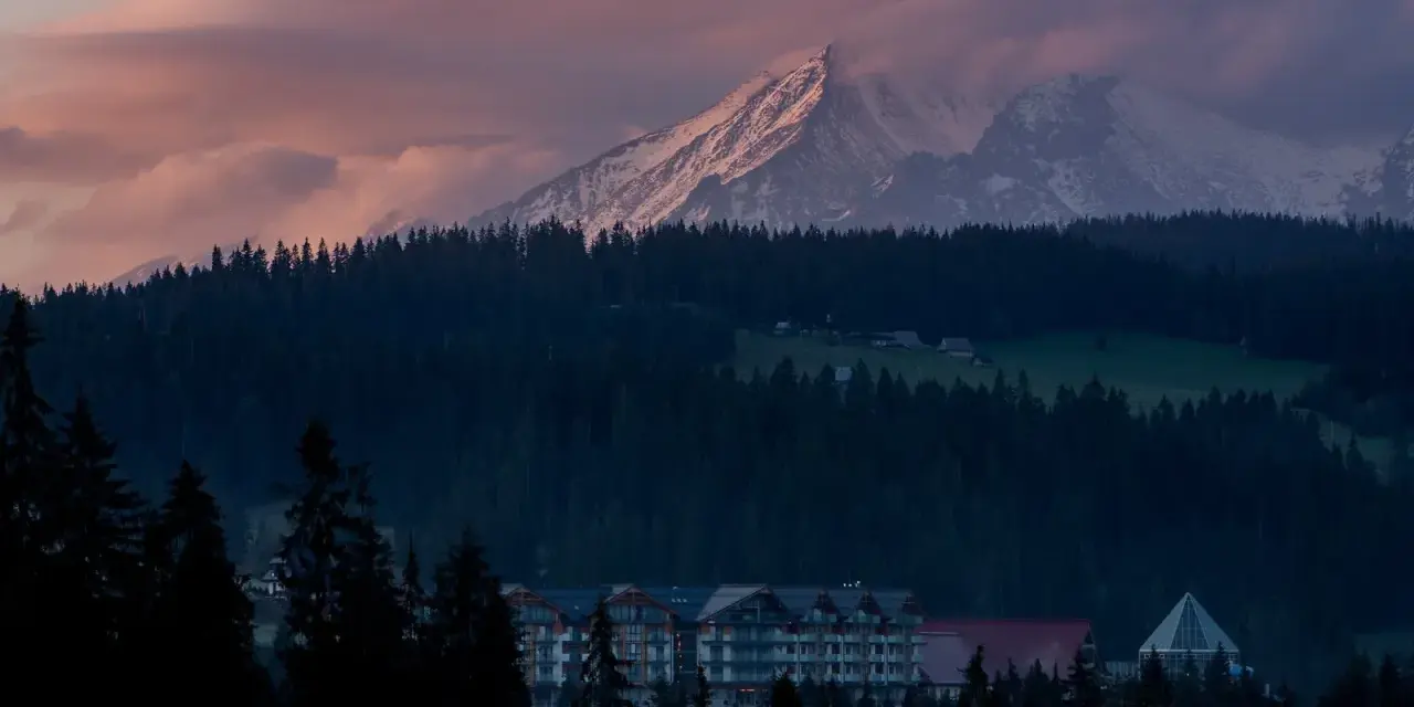 Górski krajobraz o zachodzie słońca z ośnieżonymi szczytami, lasem i budynkami hotelowymi.