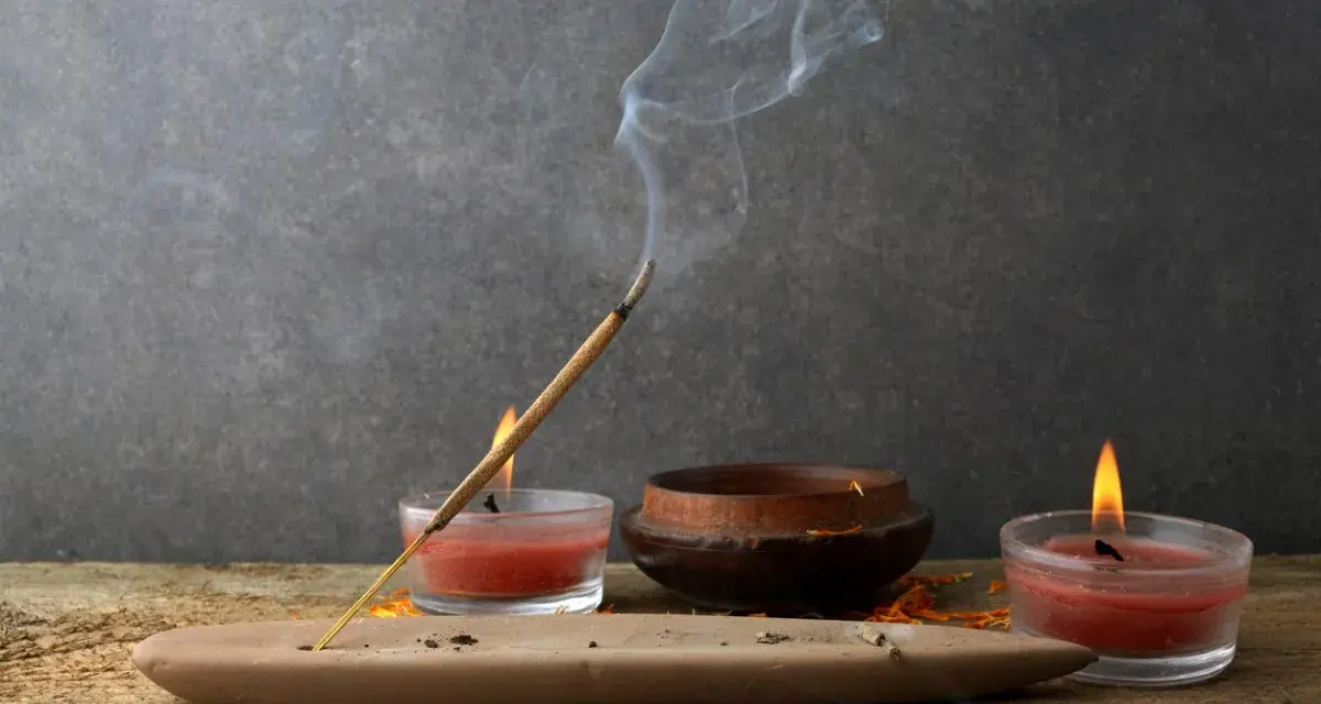 Un incienso humeante se posa sobre un soporte de arcilla, rodeado de velas rojas y un cuenco de madera. Perfectos inciensos para limpiar energías y crear un ambiente de paz.