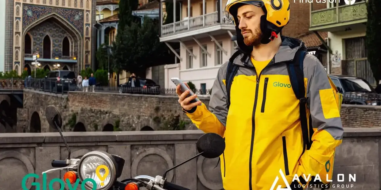 Kurier Glovo sprawdza telefon, gotowy do pracy w blokach czasowych. Na skuterze widnieje logo.