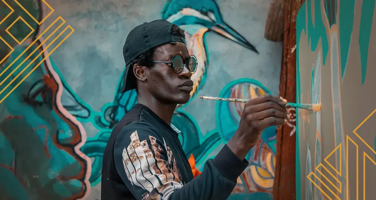 Un artiste au style d'art peinture, casquette et lunettes rondes, peint une fresque murale colorée représentant un visage et un oiseau.
