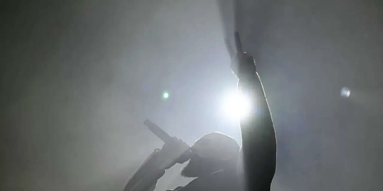 Un rappeur, micro en main, lève un doigt en signe de paix. Lumière intense, fumée, ambiance d'un exposé sur le rap.