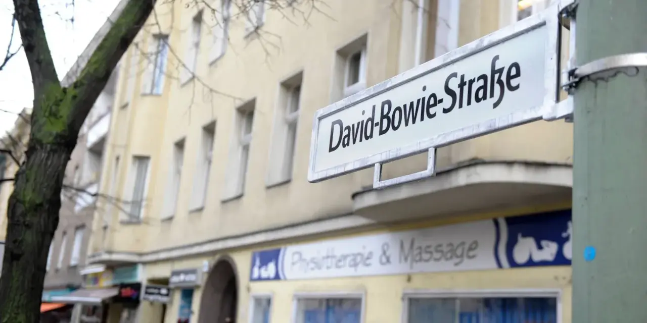 Straßenschild "David-Bowie-Straße" in Berlin. Ein Baum und ein Gebäude mit Geschäften im Hintergrund.