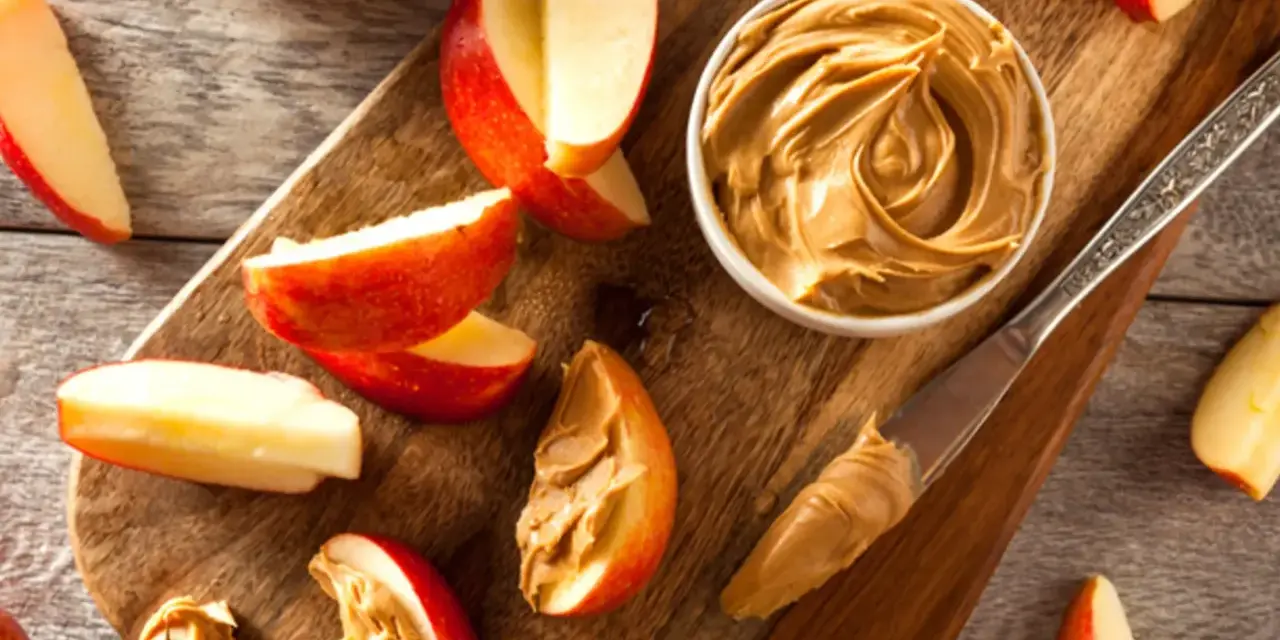 Saftige Apfelspalten mit cremiger Erdnussbutter auf einem Holzbrett. Ein Messer mit Erdnussbutter liegt daneben.