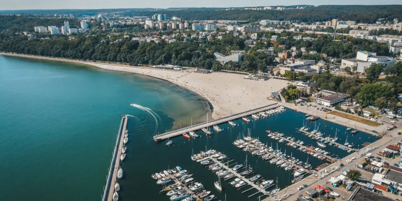 Widok z lotu ptaka na plażę miejską w Gdyni, z zatoką pełną jachtów i łodzi, oraz miastem w tle.