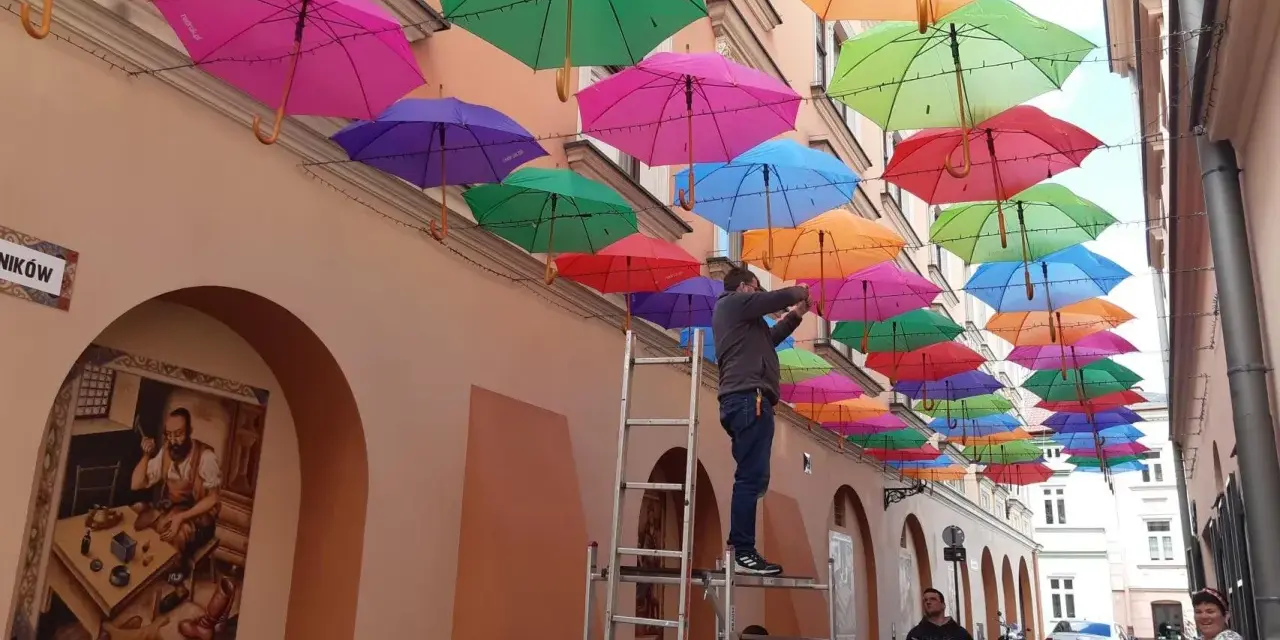 Kolorowe parasolki tworzą tęczowy dach nad uliczką w Tarnowie. Mężczyzna na drabinie dokańcza instalację.