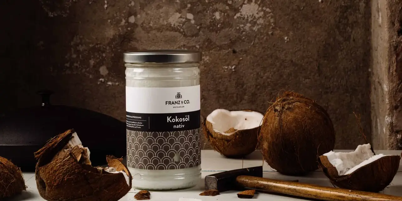 Ein Glas Kokosöl nativ steht neben zerbrochenen Kokosnüssen und einem Hammer. So wird aus der Frucht das wertvolle Öl gewonnen.