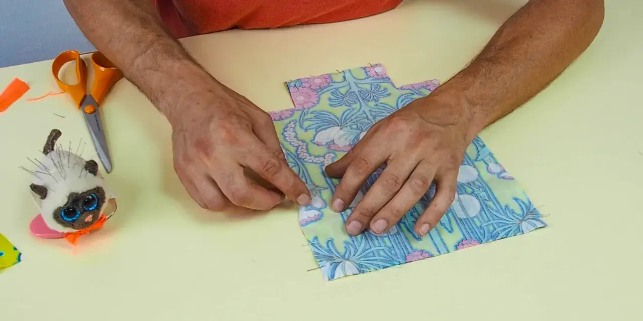 Mains assemblant deux pièces de tissu fleuri, parfait pour une idée couture débutant. Ciseaux et un chat porte-épingles à proximité.