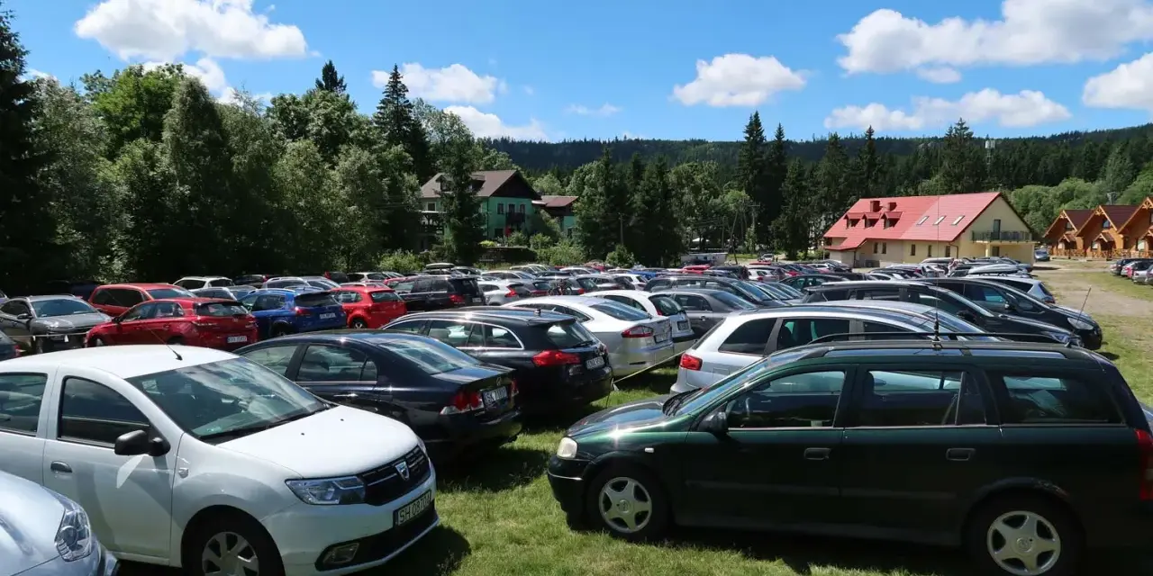 Tłum samochodów na parkingu przy Karłowie Szczeliniec. Słoneczny dzień, chmury na niebie, otoczenie lasu i budynków.