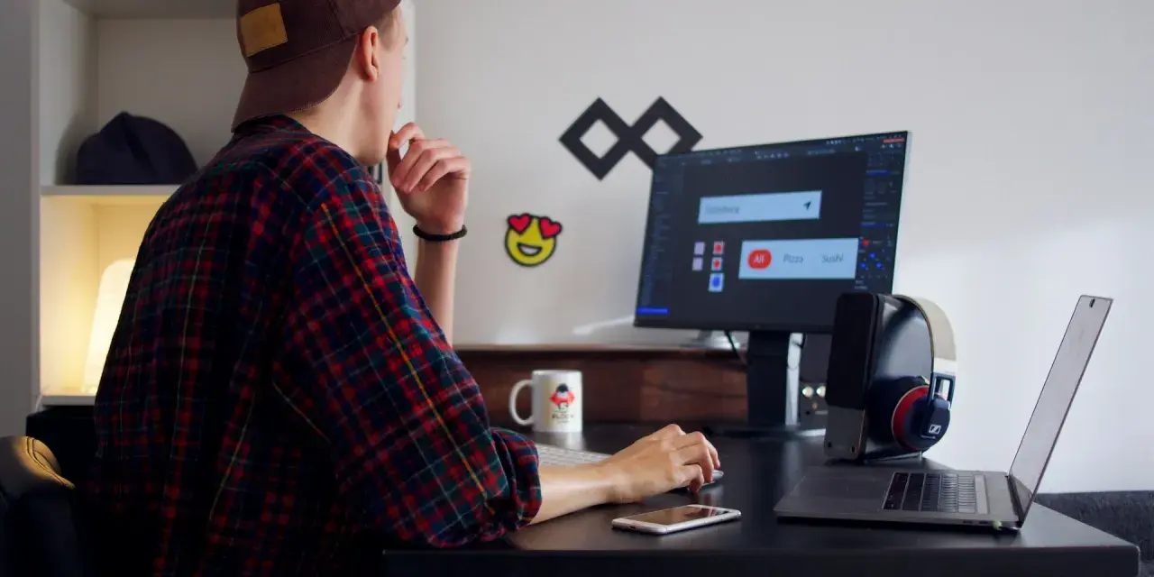 Młody mężczyzna w czapce i flanelowej koszuli pracuje przy komputerze. To typowy freelancer, który tworzy swoje projekty z domu.