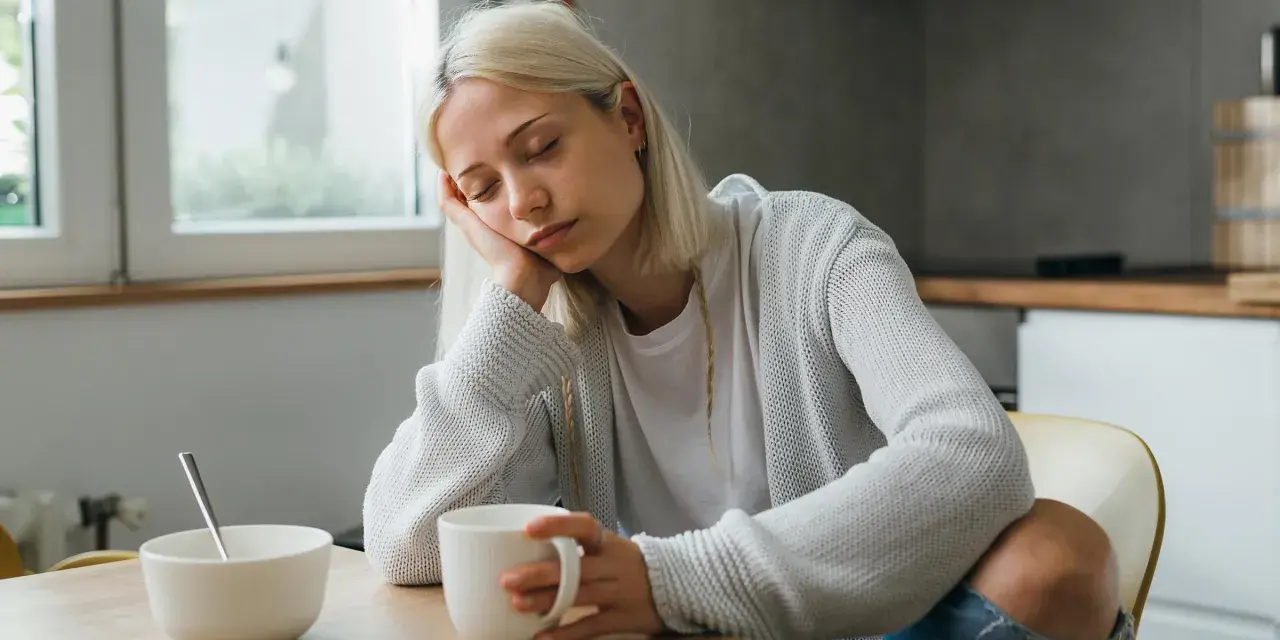 Zmęczona kobieta z blond włosami opiera głowę na dłoni, trzymając kubek. Może to być jeden z objawów keto grypy.