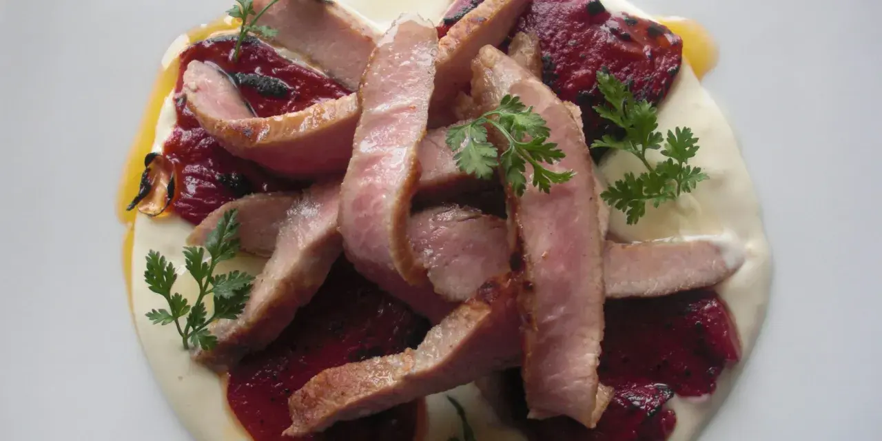 Lonchas de cerdo tierno sobre una cama de puré cremoso y pimiento del piquillo asado, decorado con perejil fresco.