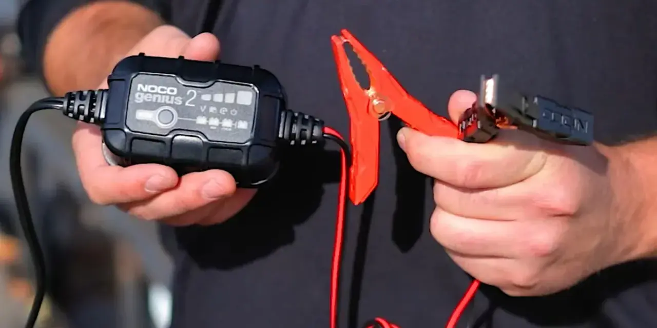 A person holds a NOCO Genius 2 trickle charger and its red clamp. This charger is designed to prevent damage, so no, a trickle charger can't ruin a battery when used correctly.