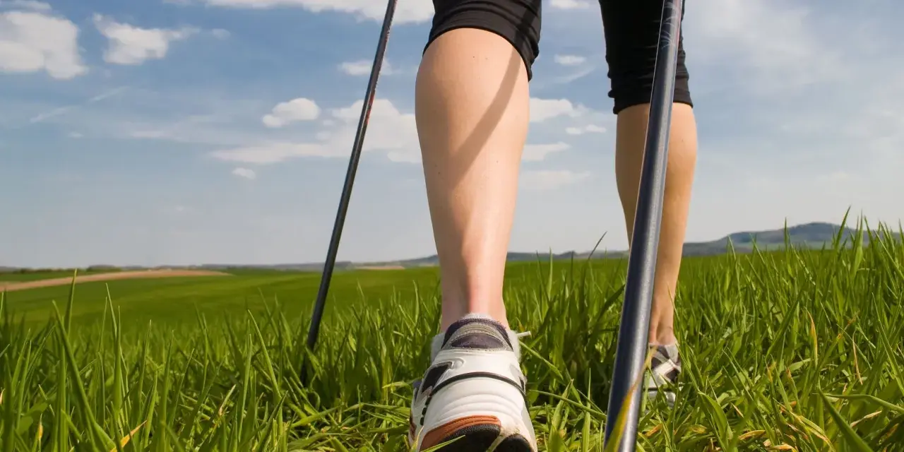 Gambe che praticano nordic walking tecnica su un prato verde sotto un cielo azzurro con nuvole bianche.