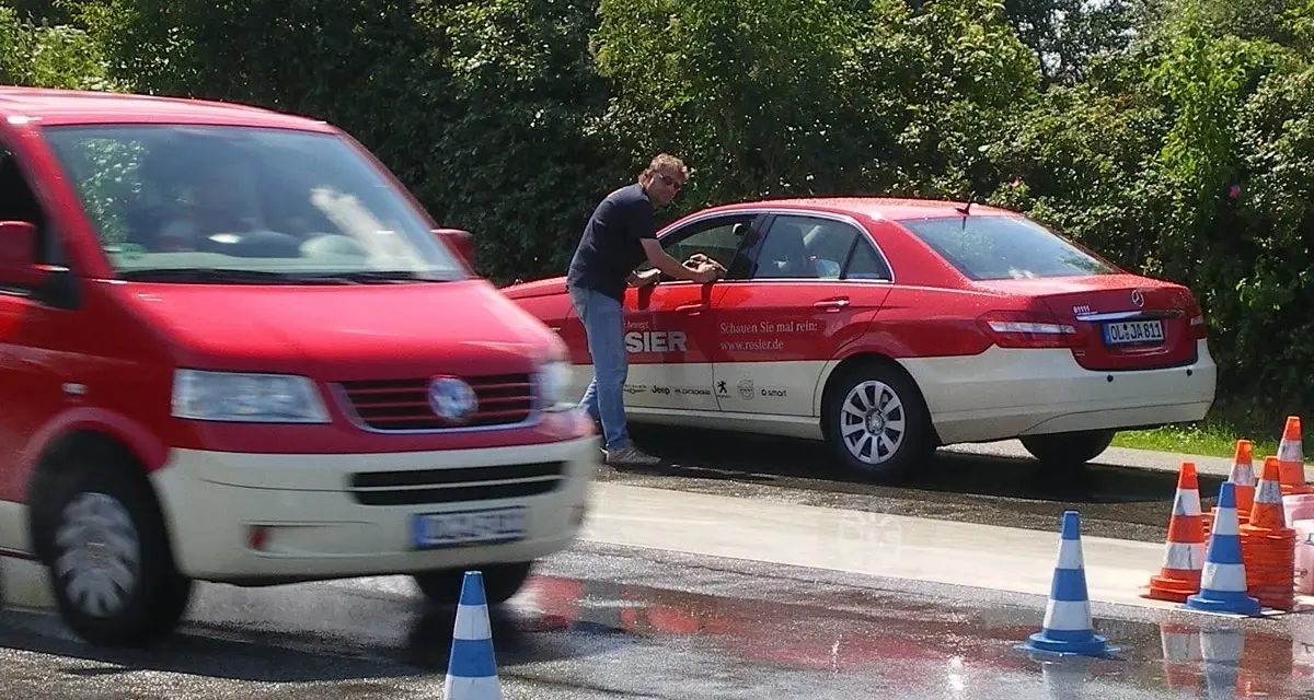Mann reinigt Taxi. Ein roter Transporter fährt vorbei. Verkehrskegel markieren den Bereich. Möglicherweise eine Schulung der Berufsgenossenschaft Taxi.