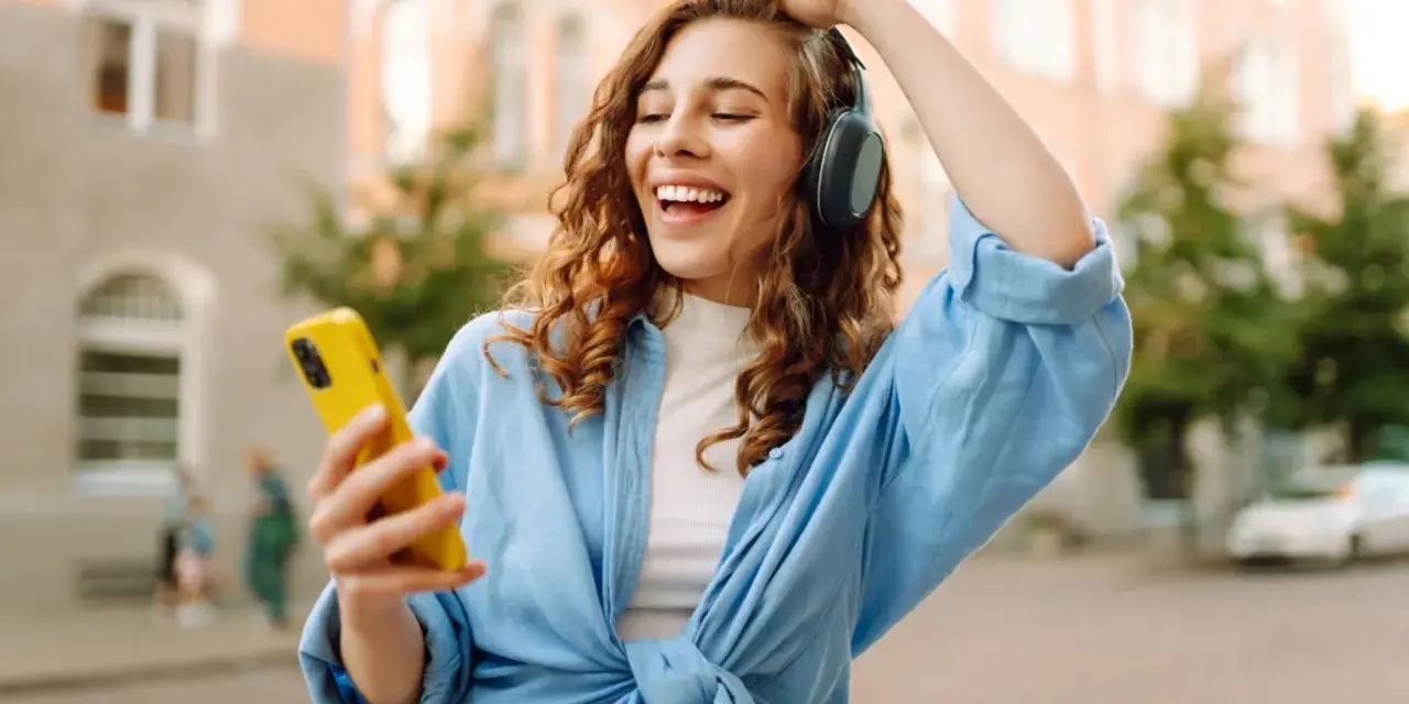 Kobieta w słuchawkach i niebieskiej koszuli trzyma żółty telefon, słuchając muzyki.