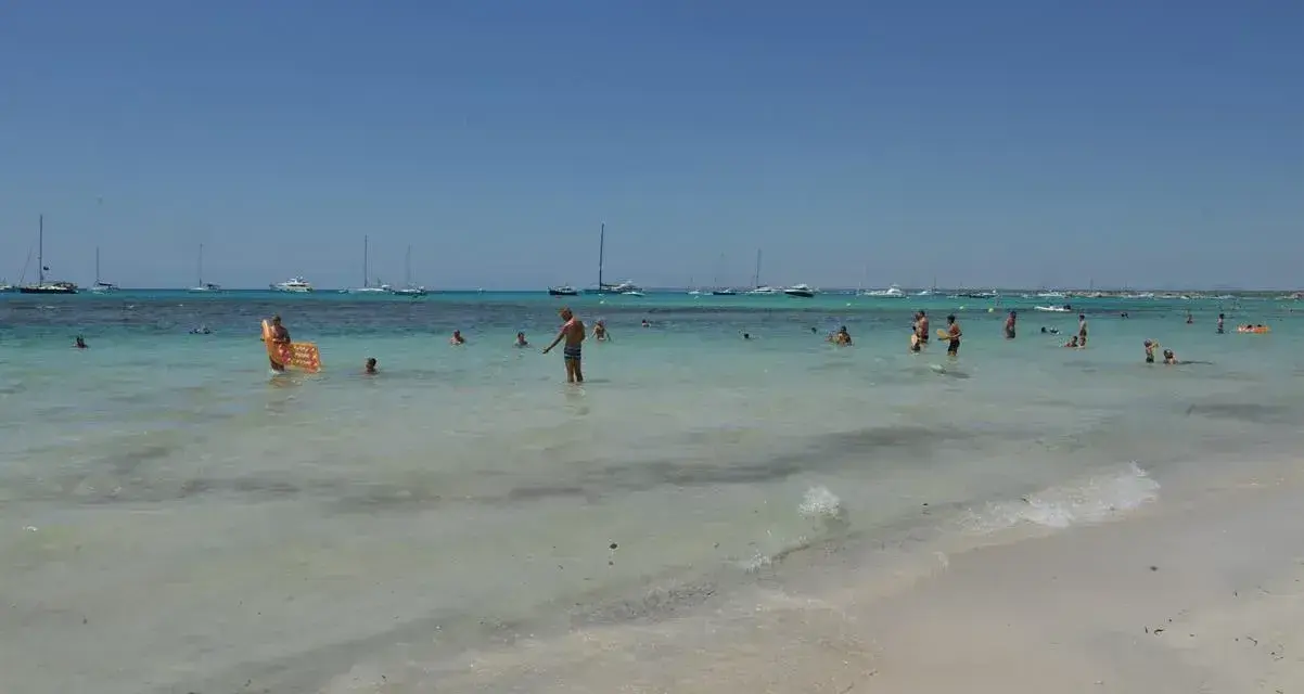 Słoneczny dzień na plaży Es Trenc. Ludzie kąpią się w turkusowej wodzie, a na horyzoncie widać żaglówki.
