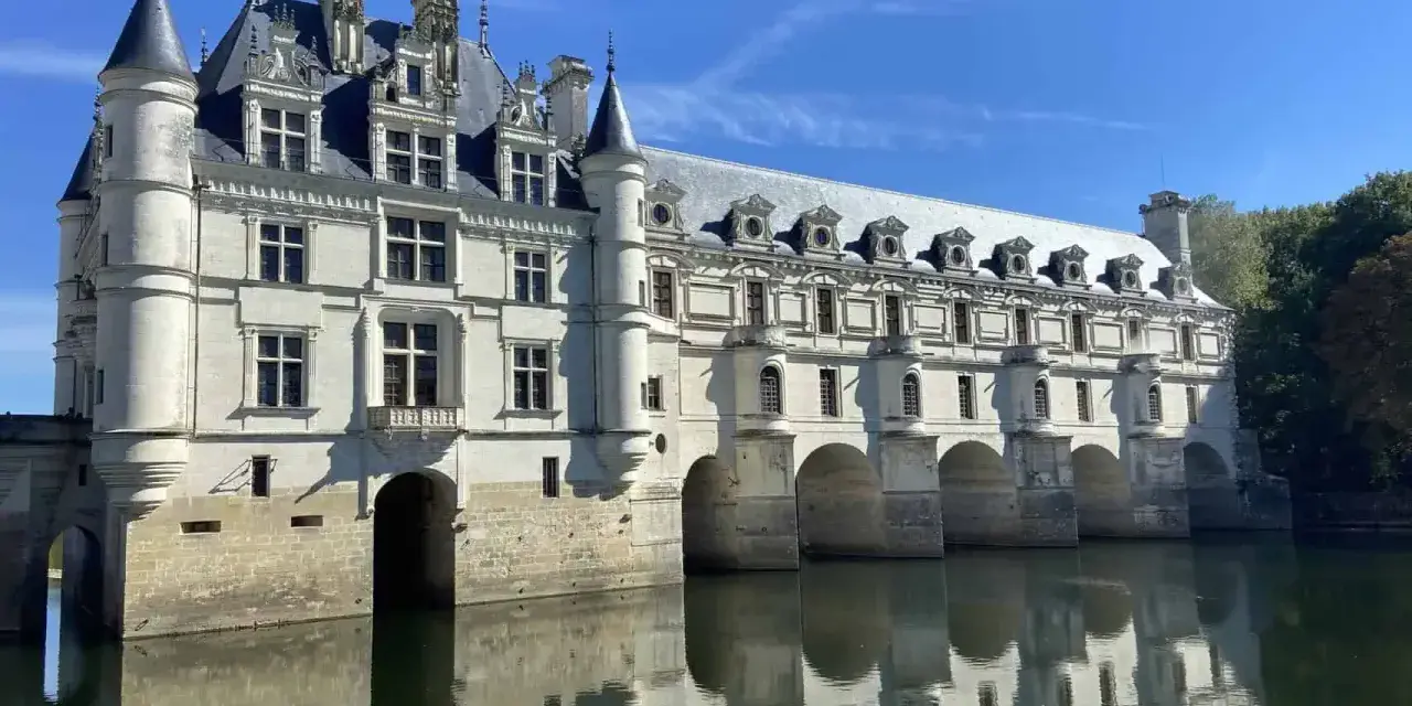 The stunning Chateau de Chenonceau, a white stone castle, gracefully spans the River Cher, its reflection shimmering on the water's surface under a clear blue sky.