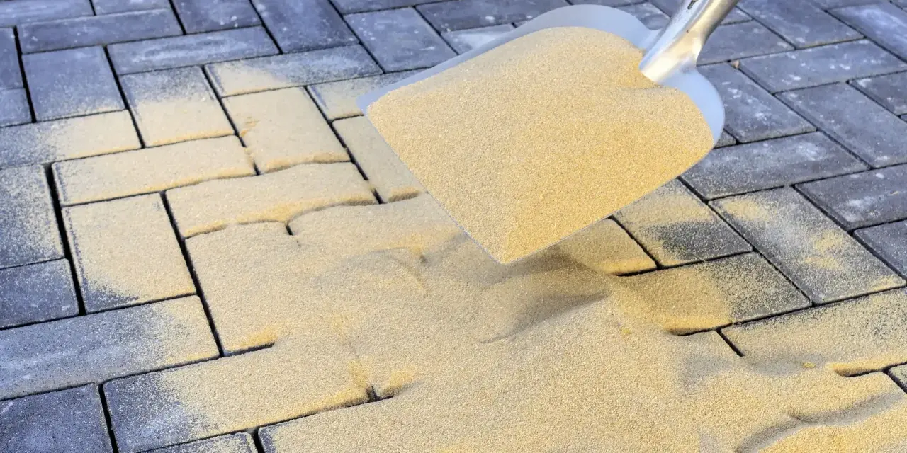 Schaufel mit Sand zum Terrassenplatten verfugen. Sand rieselt auf graue Pflastersteine.