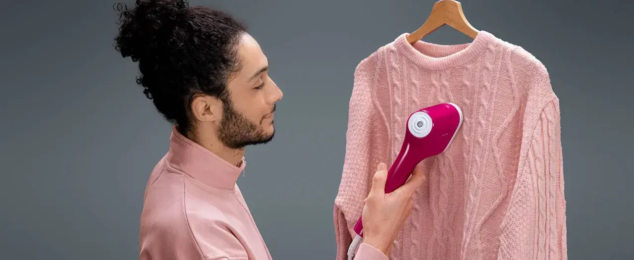 Man using a pink handheld steamer on a pink cable-knit sweater hanging on a wooden hanger.
