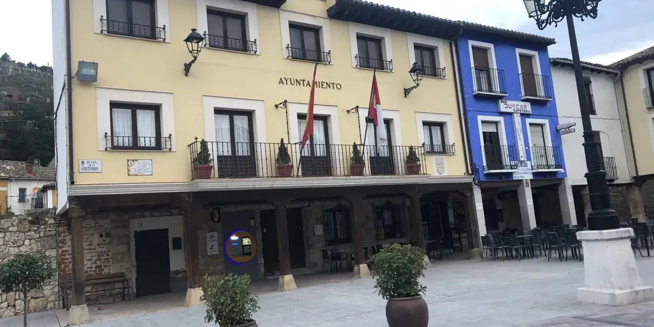 El Ayuntamiento de Tajuña, con su balcón y reloj, preside la plaza. Edificios de colores y farolas completan la escena.
