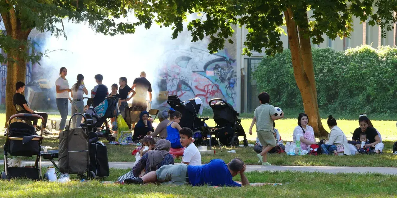 Familien genießen den Sommer auf einem Grillplatz in Berlin. Rauch steigt von einem Grill auf, während Kinder spielen und sich entspannen.