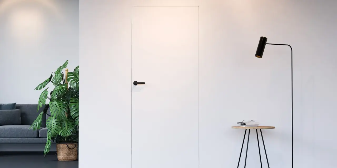 Białe drzwi ukryte w ścianie z czarną klamką, obok stojąca lampa i stolik.