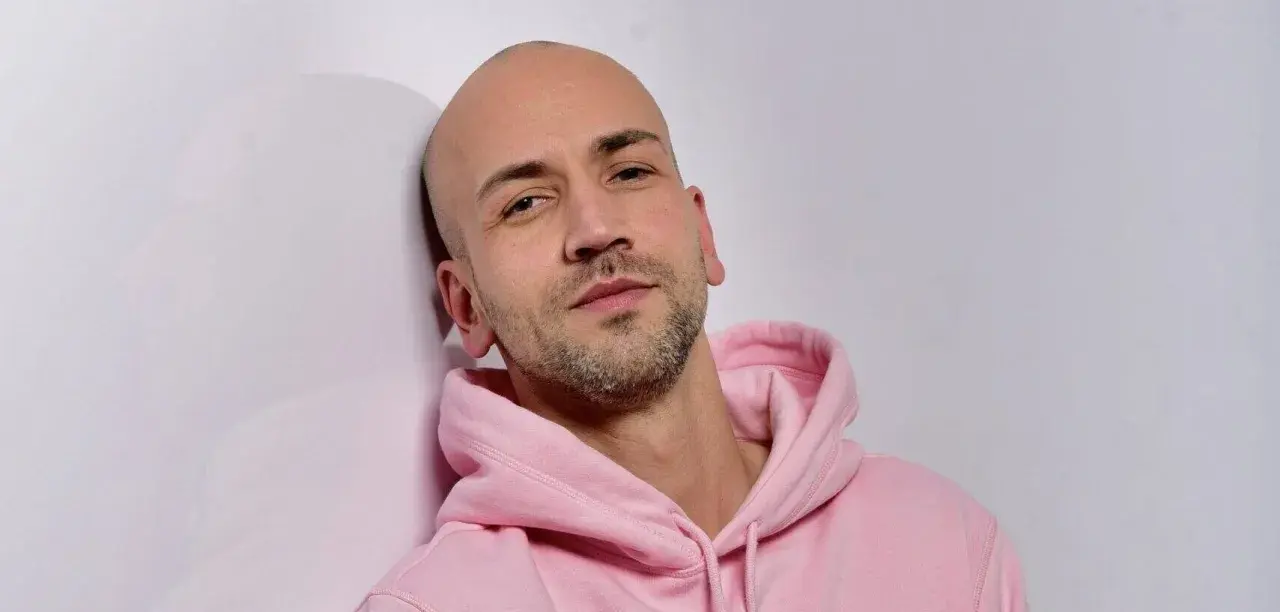 Ein Mann mit Glatze und Bart trägt einen rosafarbenen Kapuzenpullover und lehnt an einer weißen Wand.