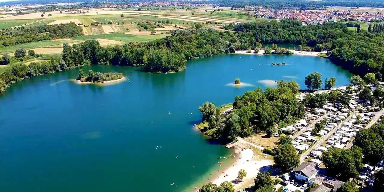 Der Mechtersheim See mit seiner türkisen Wasserfarbe, umgeben von grünen Bäumen und einem Campingplatz. Im Hintergrund erstrecken sich Felder und eine Stadt.