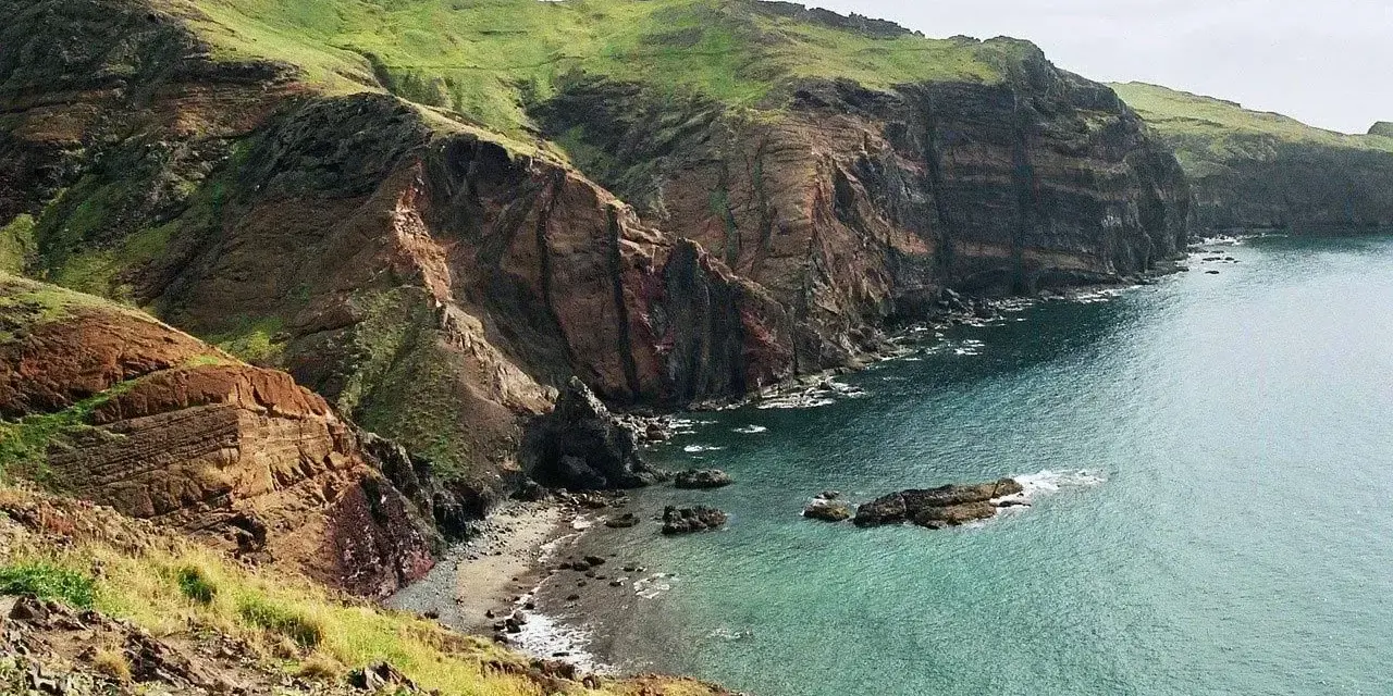 Wysokie, zielone klify Madery w sierpniu, z turkusową wodą oceanu obijającą się o skaliste wybrzeże.