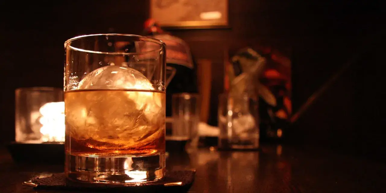 Ein Glas mit bernsteinfarbenem Whisky und einer großen Eiskugel steht auf einem dunklen Holztisch. Man trinkt Whisky mit Eis für den perfekten Genuss.