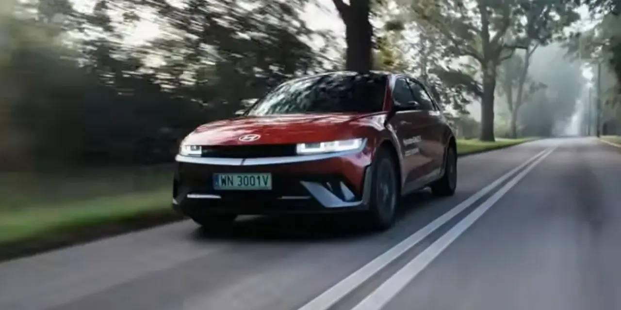 Czerwony Hyundai Ioniq 5 jedzie po wiejskiej drodze wśród drzew.
