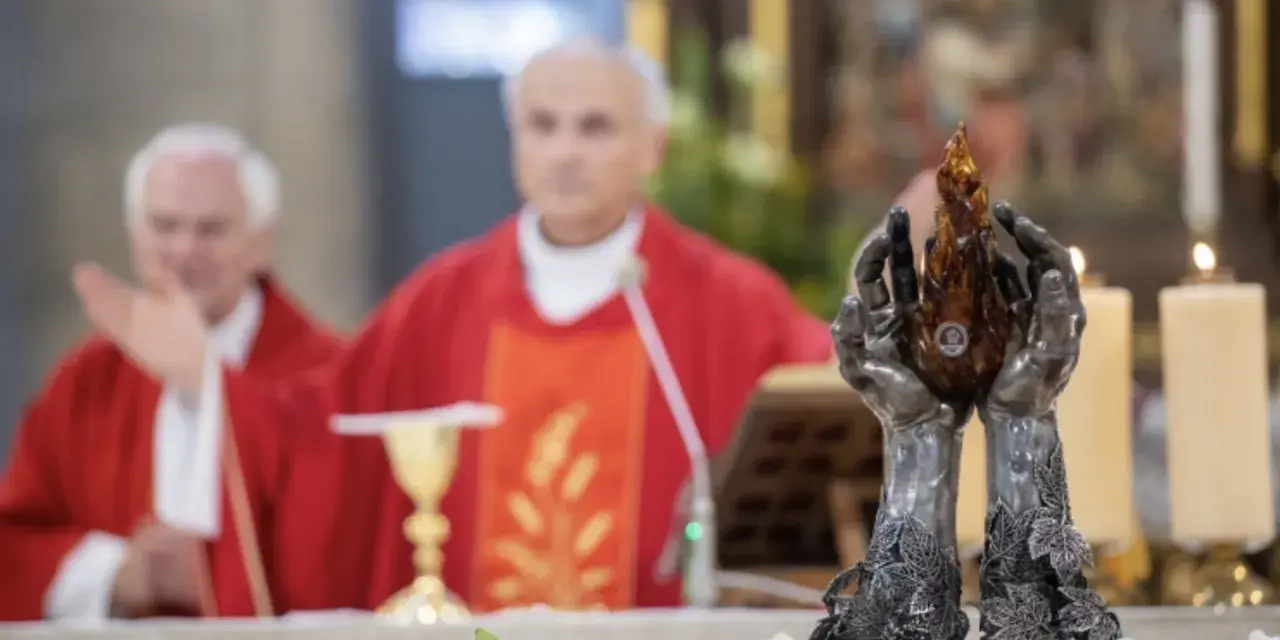 Dwie dłonie trzymają płomień. W tle ksiądz w czerwonej szacie, jak siostra zakonna, błogosławi wiernych.