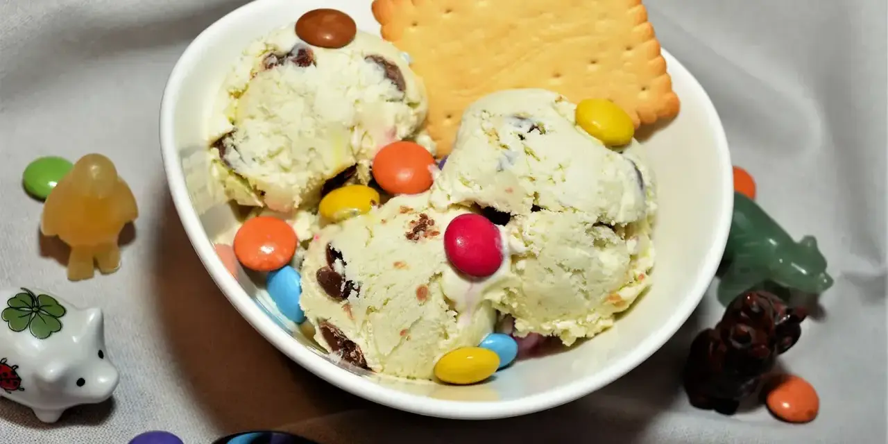 Schüssel mit cremigem Eis, garniert mit bunten Smarties und einem Keks. Kleine Figuren schmücken die Szene.