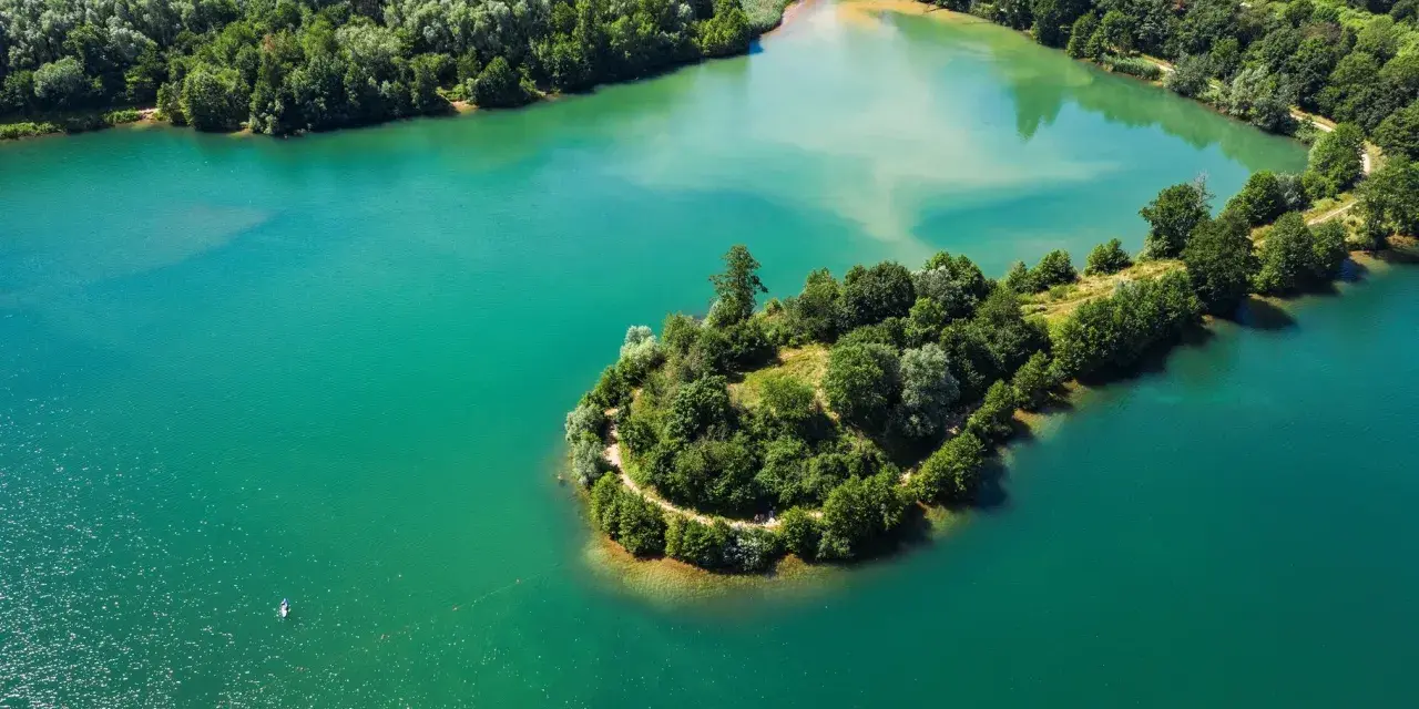 Türkisblaues Wasser umgibt eine grüne Insel mit einem Pfad am Niedernberger See. Ein Stand-Up-Paddler genießt die Ruhe.