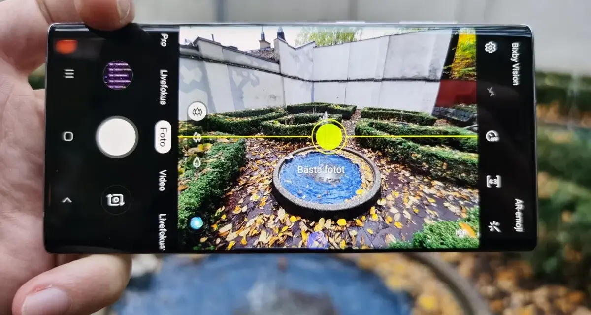 En hand håller en Samsung Galaxy Note 10 Plus med kameran öppen, redo att ta en bild av en fontän i en höstträdgård.