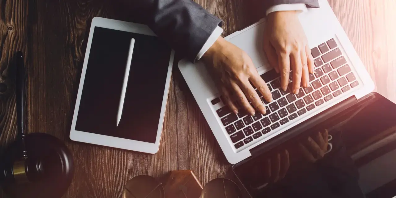 Dłonie na klawiaturze laptopa, obok tablet i młotek sędziowski. Profesjonalne narzędzia do pracy, być może związane z **taxat windykacja**.