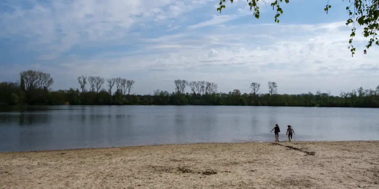 Zwei Kinder planschen im Wasser am Sandstrand des Dieksees in Lingen. Bäume säumen das Ufer unter einem bewölkten Himmel.