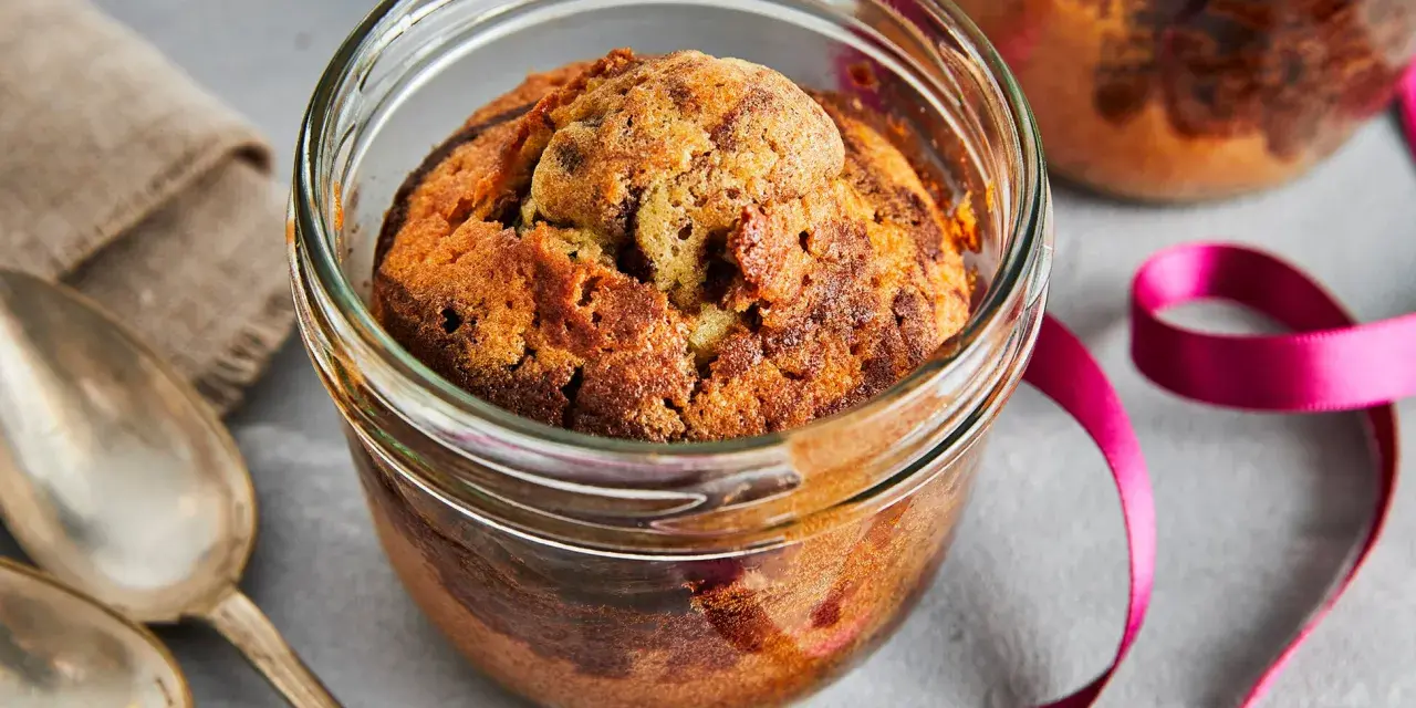 Zwei köstliche Kuchen im Glas, mit Schokoladenstückchen, warten auf Löffel. Eine pinke Schleife und Leinentuch deuten auf ein besonderes Geschenk hin.