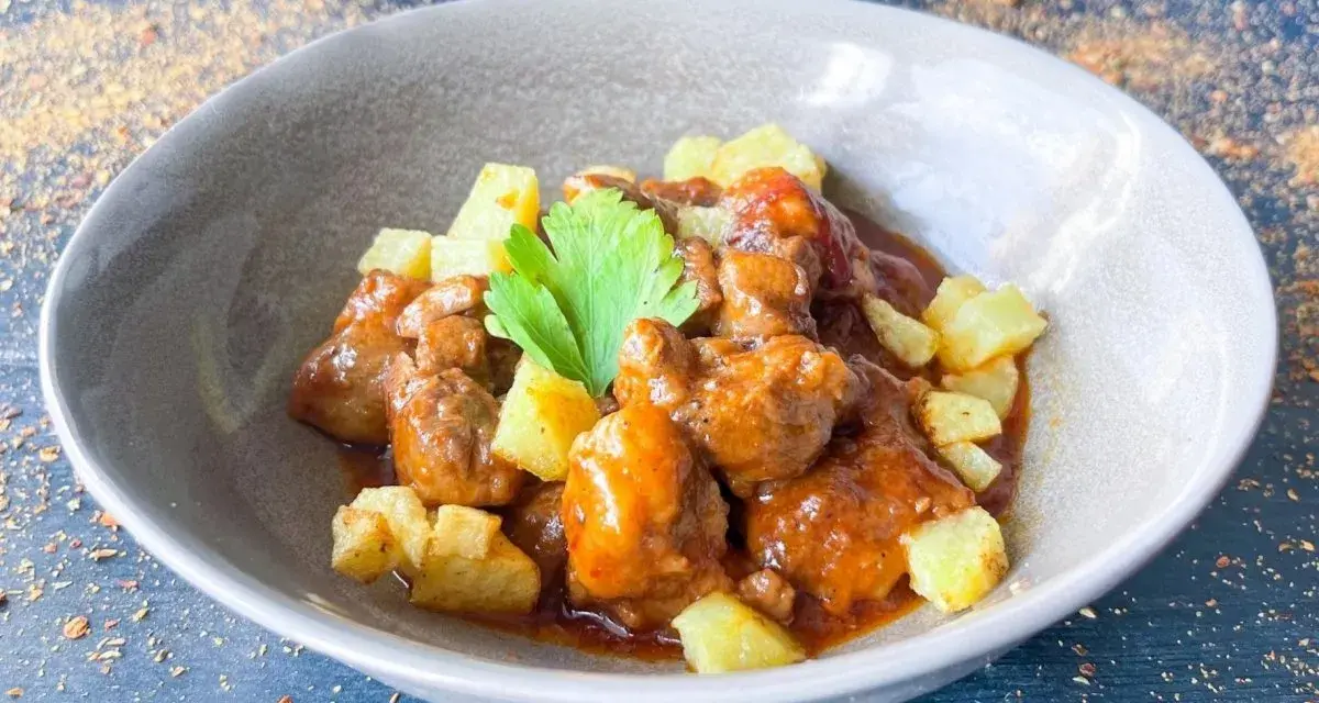 Delicioso cordero al chilindrón en un cuenco gris, acompañado de patatas fritas y una ramita de perejil.