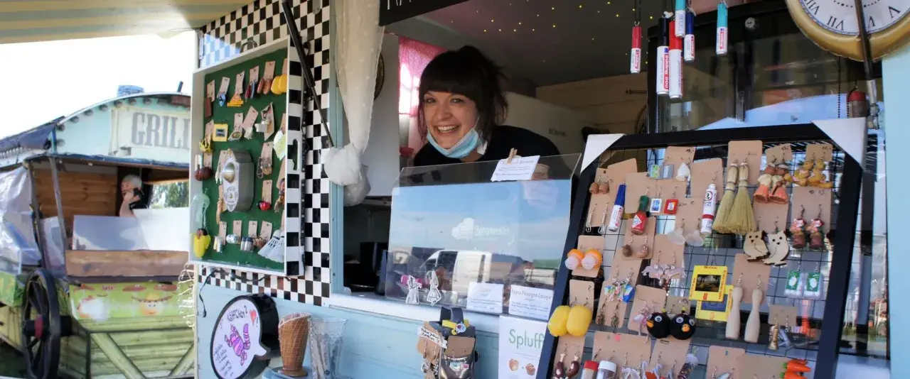 Lachende Verkäuferin auf dem Flohmarkt Friedrichshagen präsentiert handgefertigten Schmuck, darunter Eulen-Ohrringe und Miniatur-Flaschen.