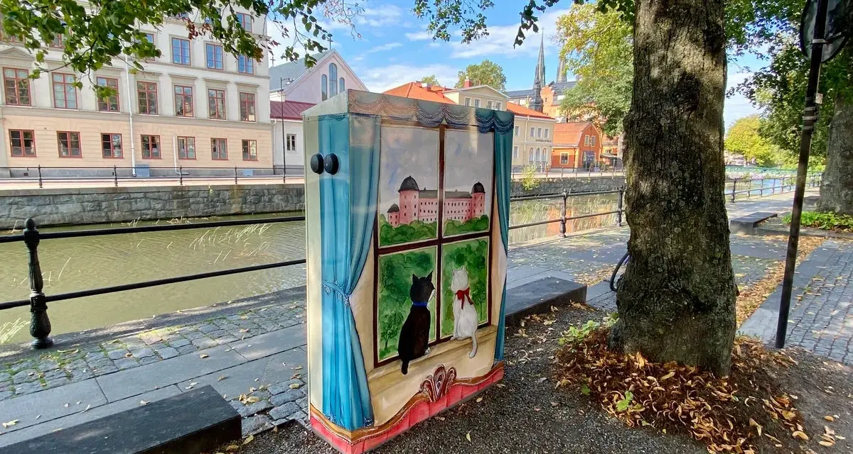 En målad låda med två katter som tittar ut genom ett fönster på ett slott. Konstnären har valt att måla tunt för att ge en drömsk känsla.