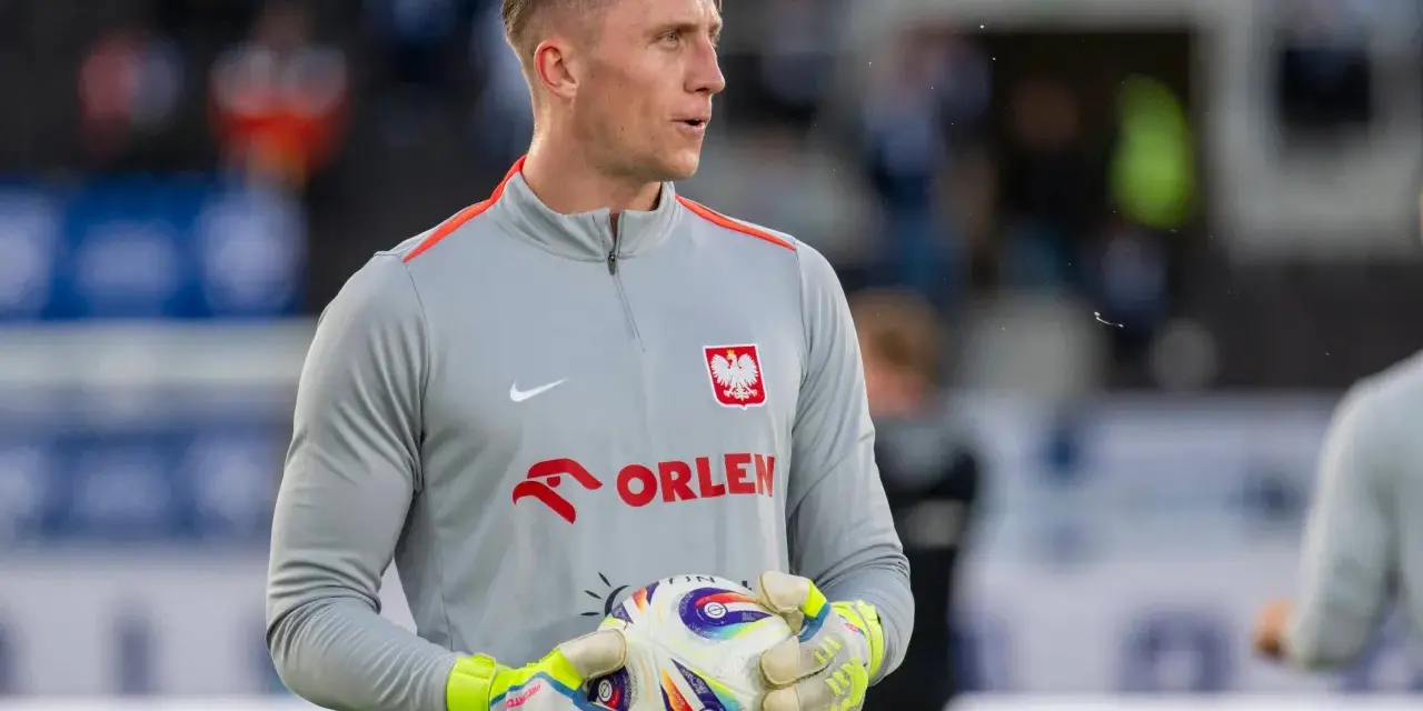 Bramkarz w szarej bluzie z orłem i napisem ORLEN, trzymający piłkę. Kolejny z polscy piłkarze za granicą, gotowy do gry.