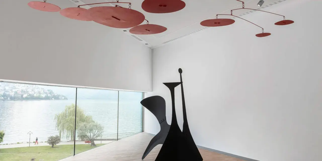 Un mobile Calder rouge flotte au-dessus d'une sculpture noire monumentale, dans une salle d'exposition lumineuse avec vue sur un lac.