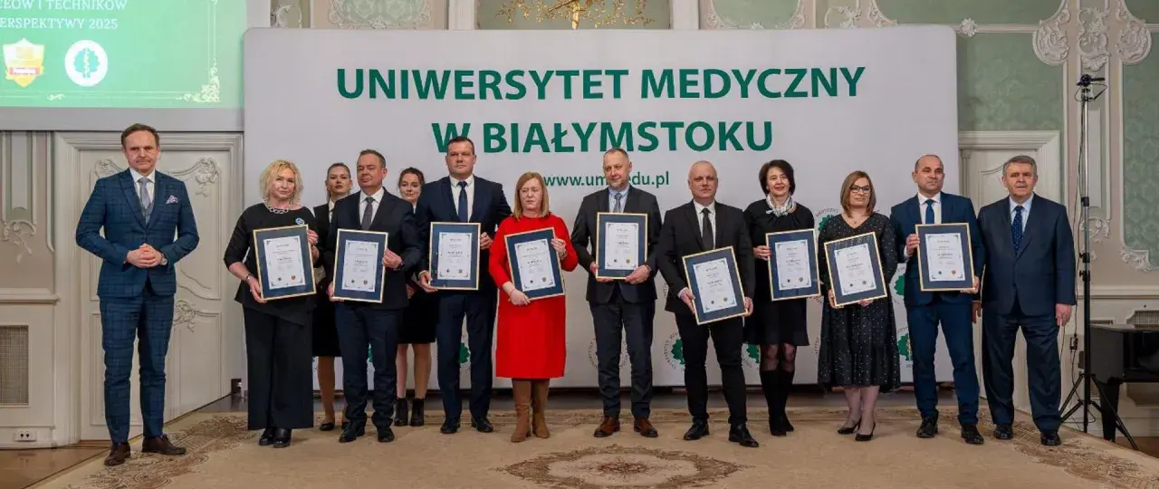 Grupa osób w garniturach i sukienkach pozuje do zdjęcia z dyplomami przed banerem Uniwersytetu Medycznego w Białymstoku.